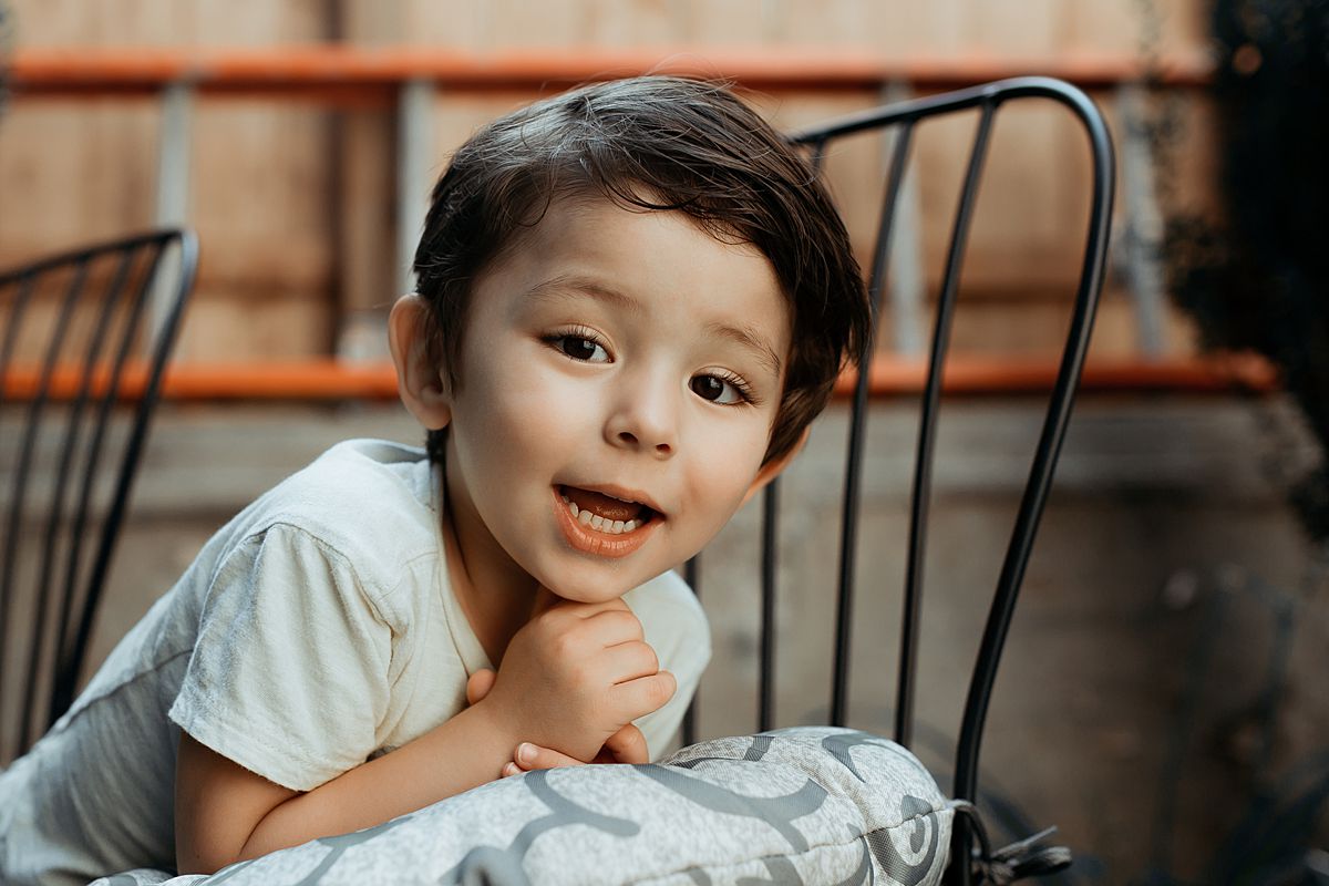 portrait of young boy posing as he leans against backyard chair. photo by nyc family and newborn photographer in harlem new york