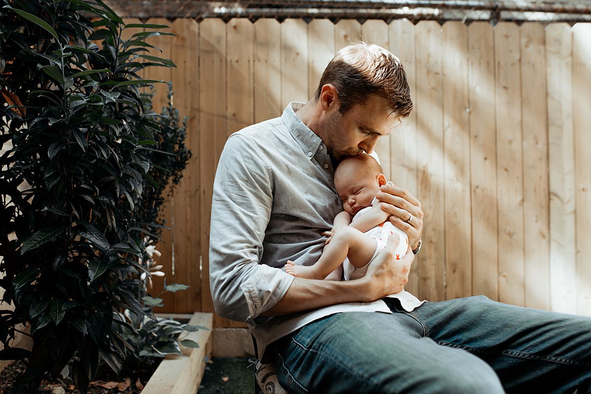 dad kisses newborn daughter while sitting outback during family lifestyle session. photo by nyc family and newborn photographer krystil mcdowall