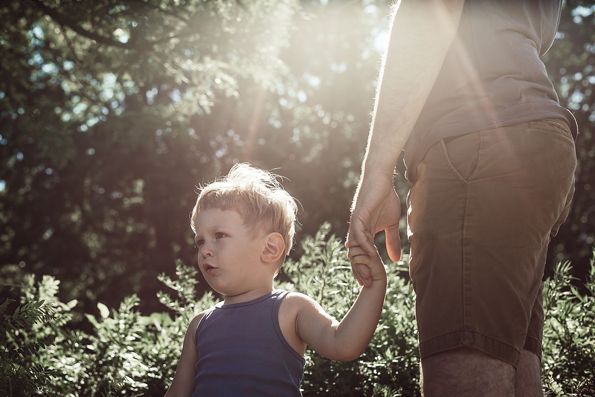 son holding dad's hand while standing in the sunshine in front of nice leafy green bushes in nyc