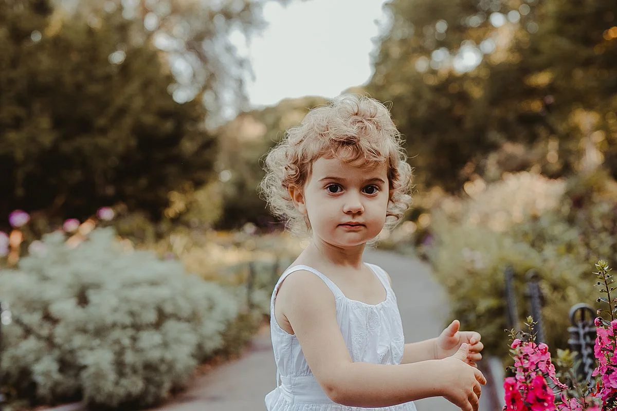 beautiful toddler girl plays amongst bright flowers in family session in fort tryon park. image by krystil mcdowall photography