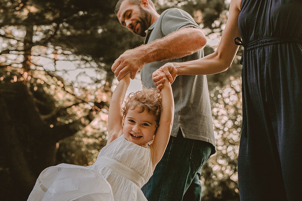 mom and dad swing daughter among fall leaves in the park during candid family session with nyc family photographer krystil mcdowall