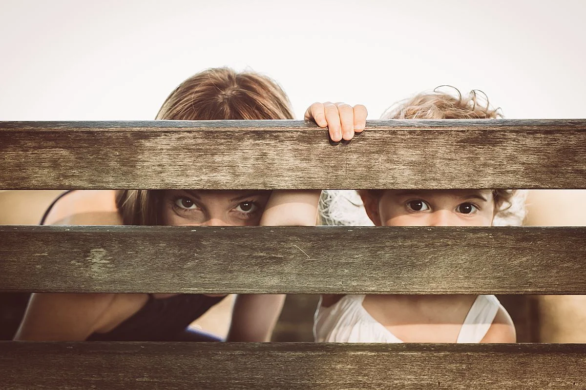 mom and daughter peek through park bench at camera. enjoying fun family moments during candid family session in nyc. photo by krystil mcdowall photography