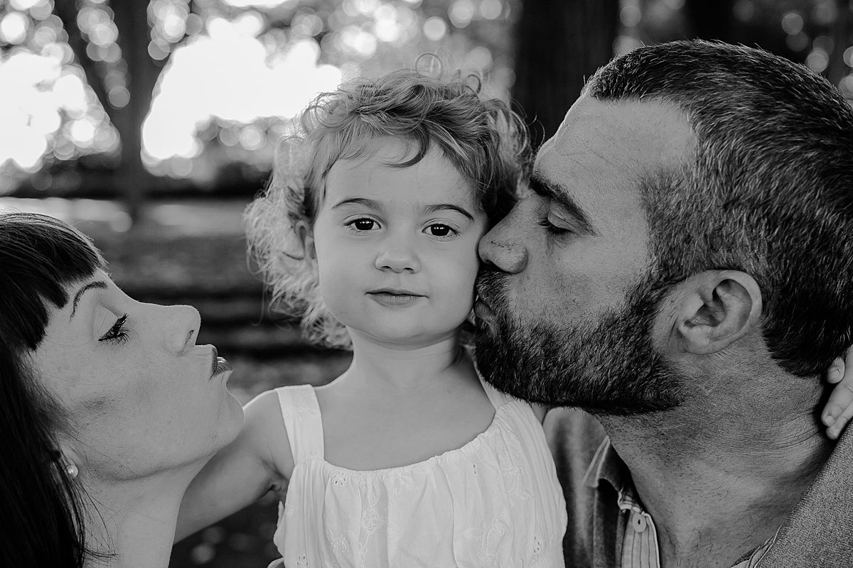 mom, dad and daughter sit on park bench squished and cuddling for kisses together. photo by krystil mcdowall