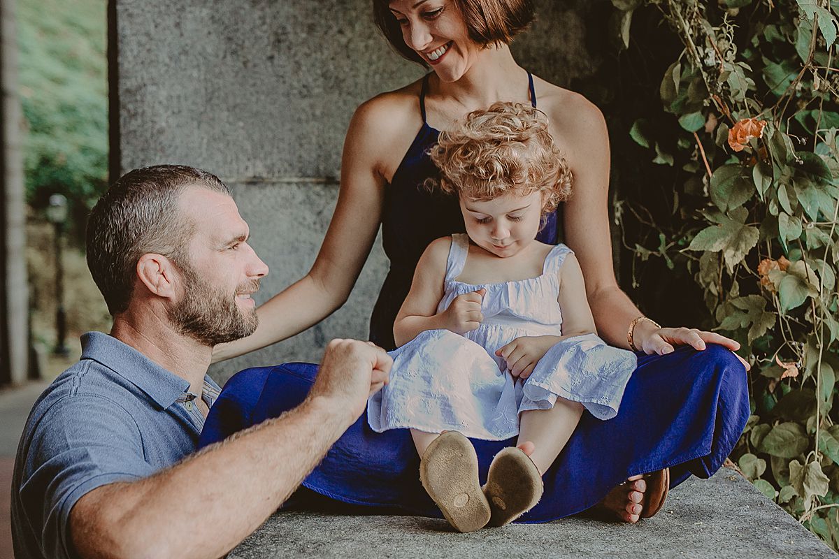 daughter sits on mom's lap with dad looking on lovingly near beautiful flower vines. photo by nyc family and newborn photographer krystil mcdowall