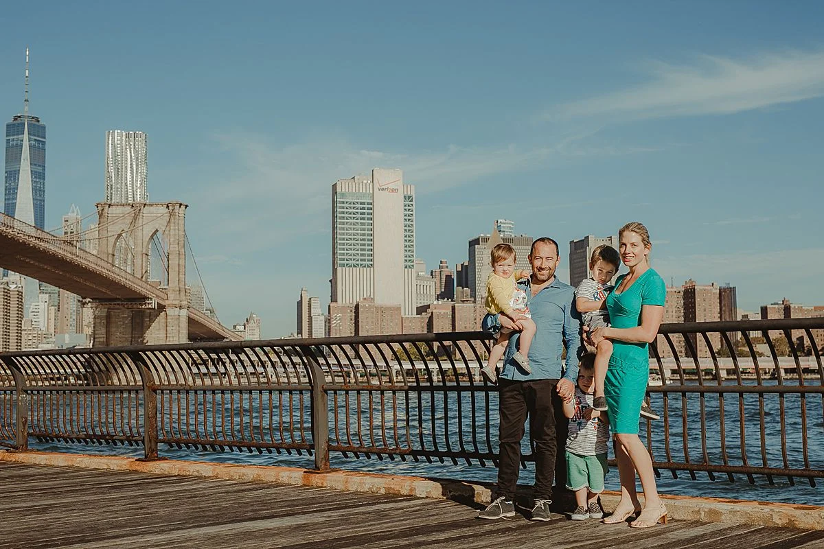 family stands on brooklyn promenade with east river and manhattan skyline in the background. photo by nyc family and newborn photographer