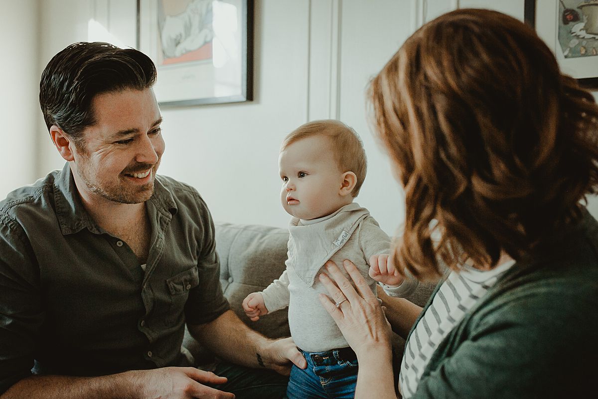Candid photo of family of three in living room of Queens nyc apartment with baby smiling and parents lovingly smiling at baby