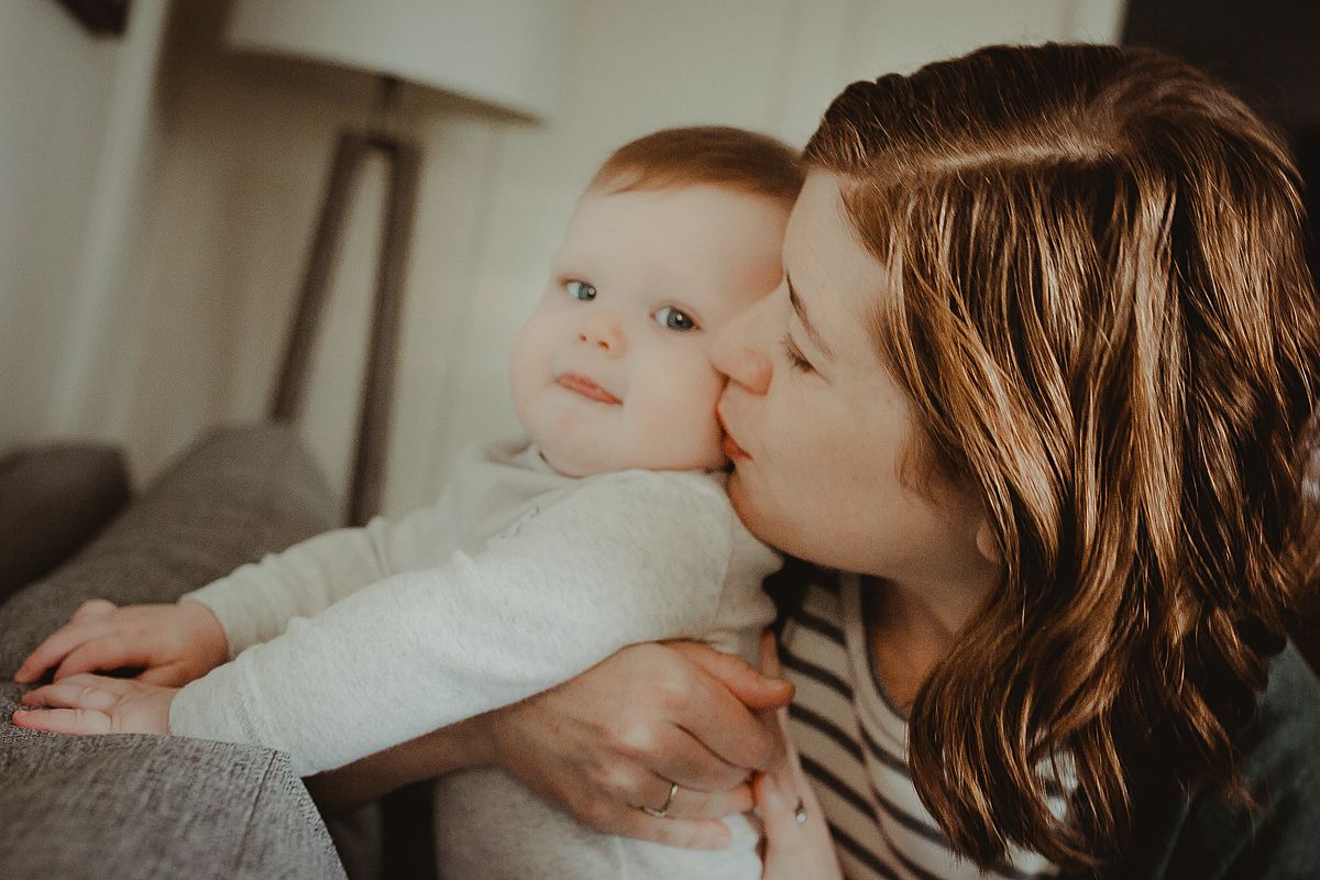 mom holds son while he explores the couch and gives son soft kiss on the cheek. krystil mcdowall photography capturing candid family photos