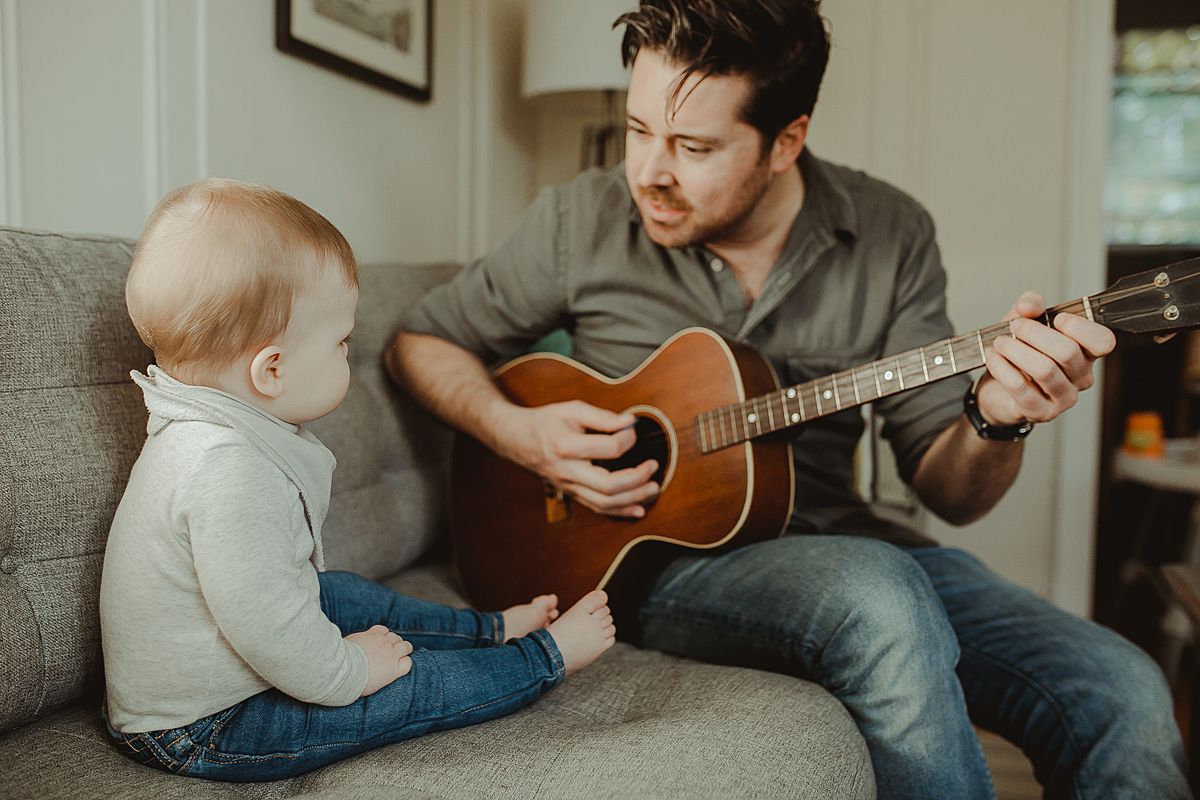 dad and son sitting on living room couch while dad plays guitar for his eight month old son during fun family photo session in queens. image by nyc family photographer krystil mcdowall