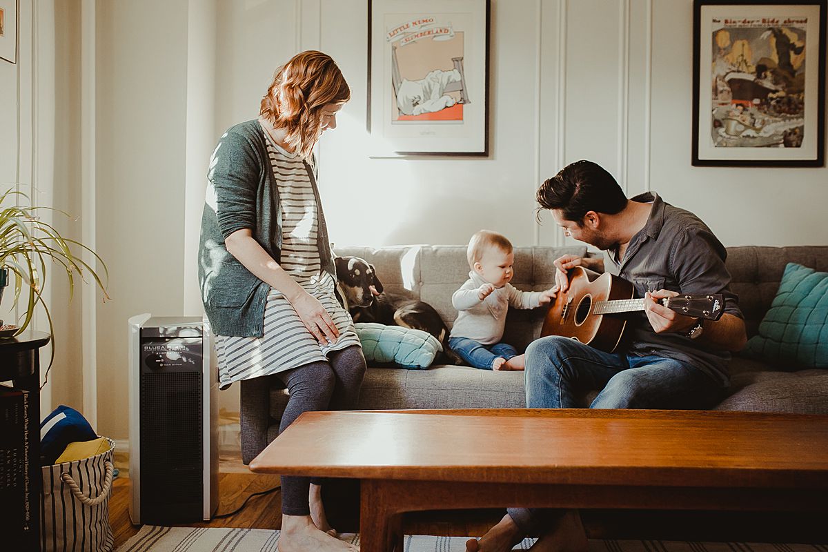 mom, baby and dog on sun filled living room couch enjoying listening to dad play guitar during documentary family photo session. image by krystil mcdowall photography