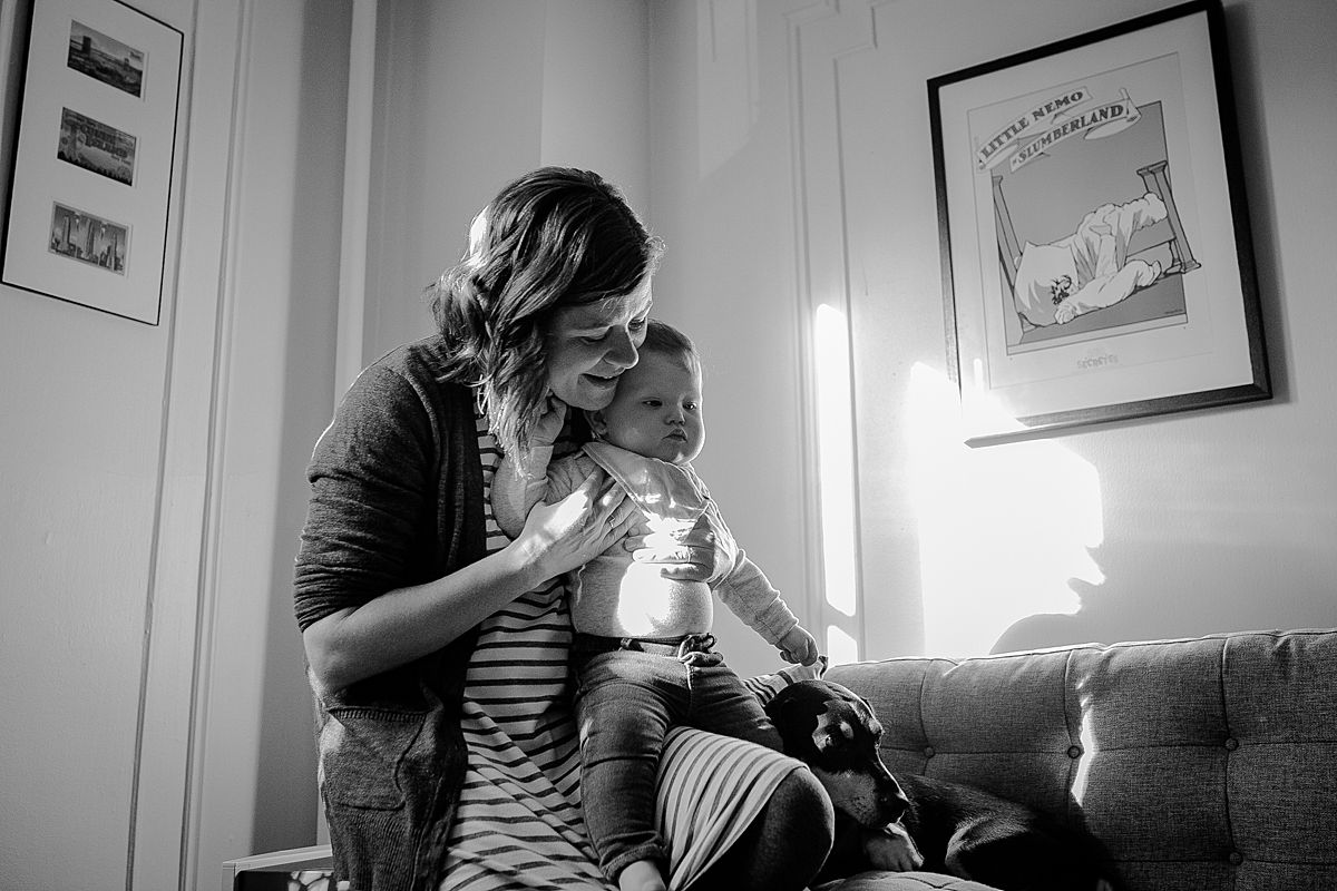 mom, baby and dog on sun filled living room couch enjoying listening to dad play guitar during documentary family photo session. image by krystil mcdowall photography