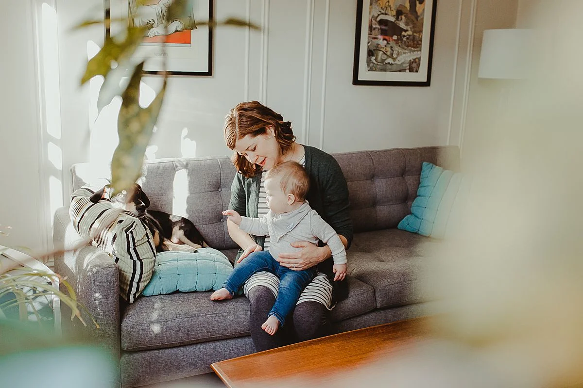 peeking at mom, baby and dog through plants in the living room during nyc family photo session. image by krystil mcdowall photography