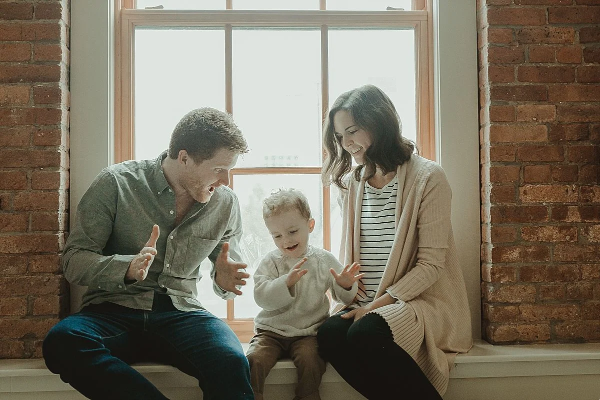 family sits in front of light filled window playing clapping hand games. krystil mcdowall ohotography capturing real candid family moments