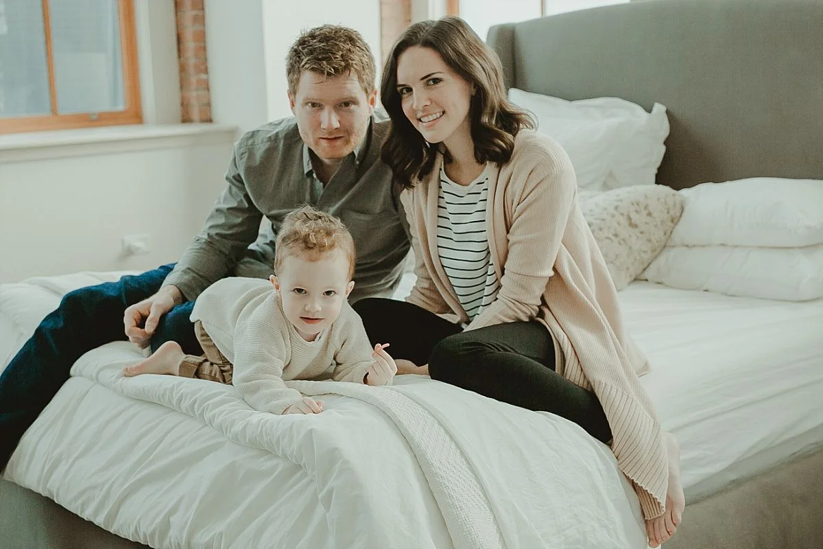 family portrait of mom, dad and son on bed and looking at camera in beautiful light filled master bedroom. photo by photographer krystil mcdowall