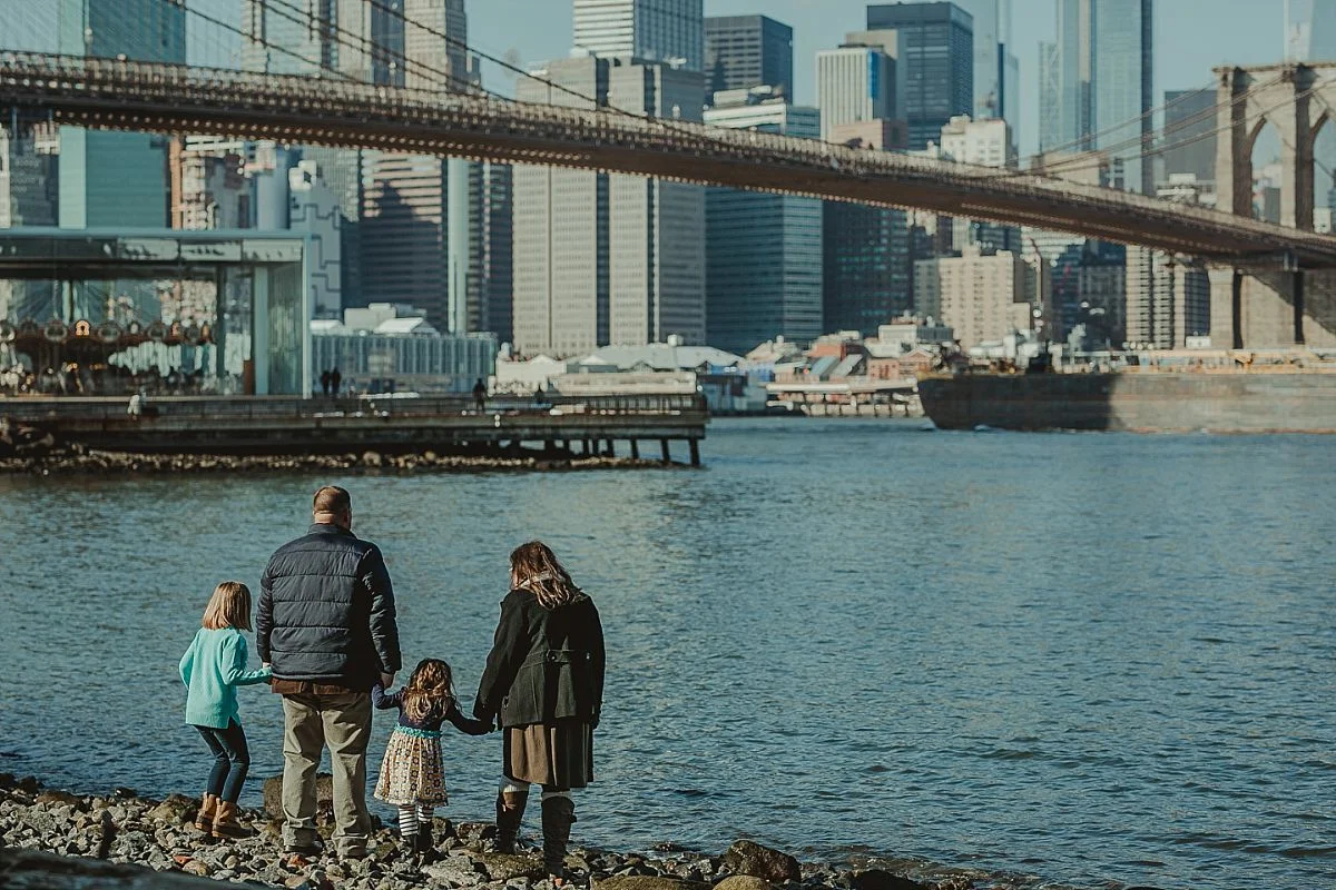 family of four at brooklyn beach holding hands as they look out at the nyc skyline. documenting real memories for your family is krystil mcdowall photography