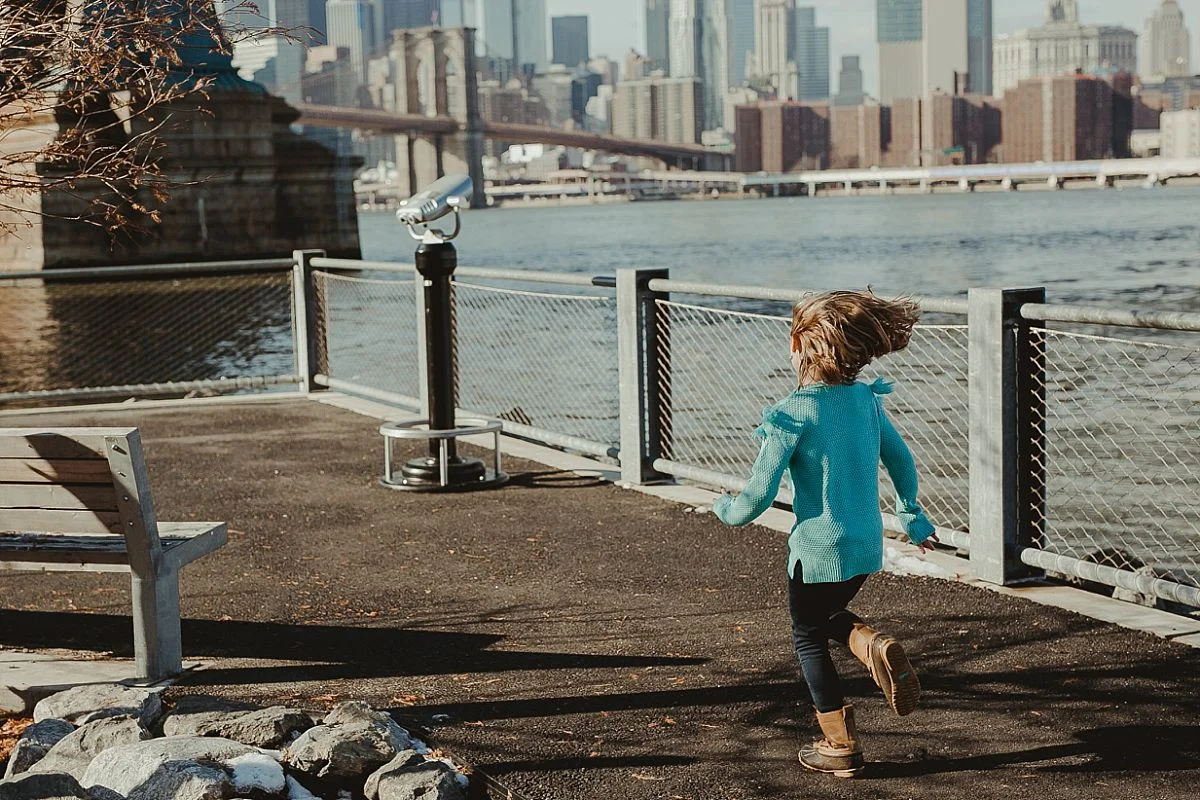 young girl in blue sweater runs along footpath in dumbo to one of the skyline lookouts located in front of the manhattan bridge. image by nyc family photographer krystil mcdowall