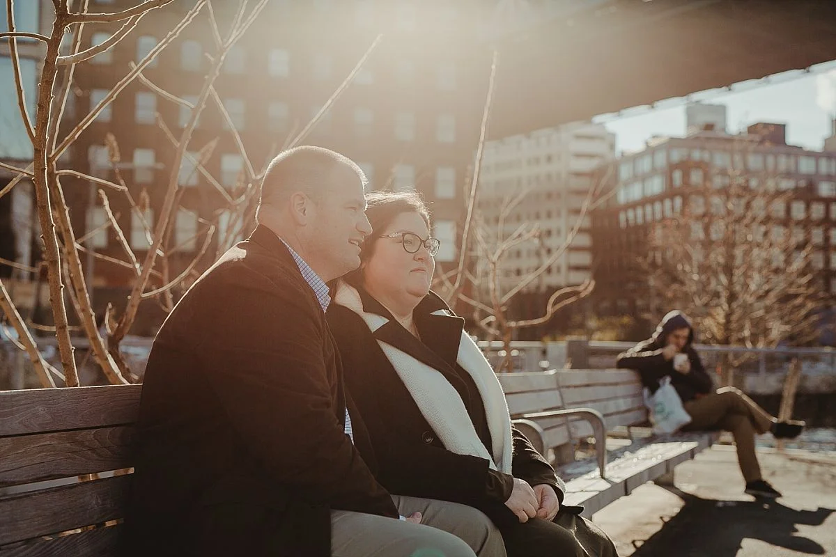photo of mom and dad taking a rest on an nyc park bench with beautiful golden sun flare in the background. image by nyc family photographer krystil mcdowall