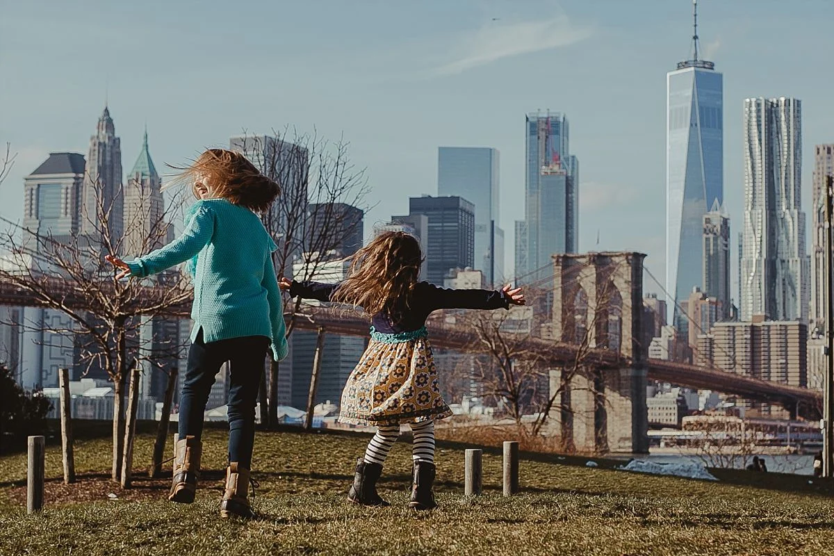 candid image of sisters dancing on green grass in brooklyn with iconic views of the nyc skyline in the background. image by nyc family photographer krystil mcdowall