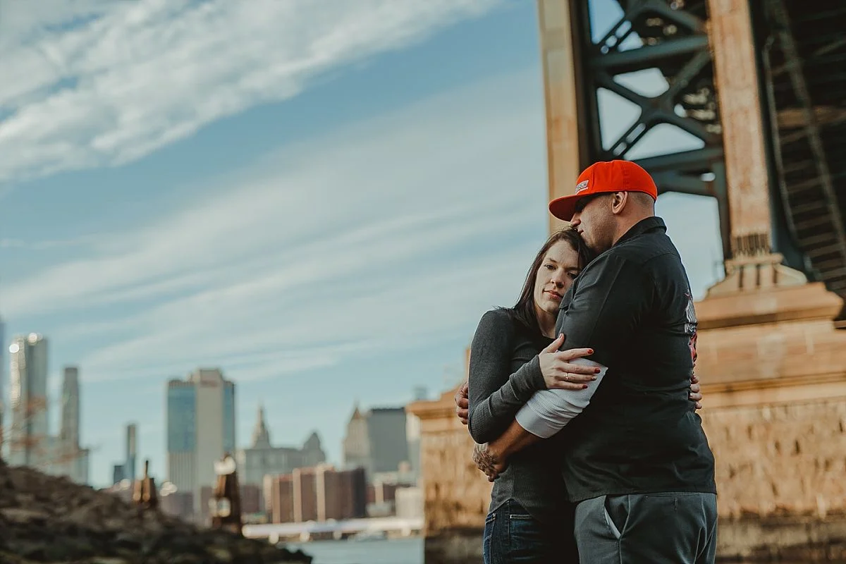 photo of couple hugging underneath manhattan bride in dumbo brooklyn during couples photoshoot. krystil mcdowall photography takes candid family photos
