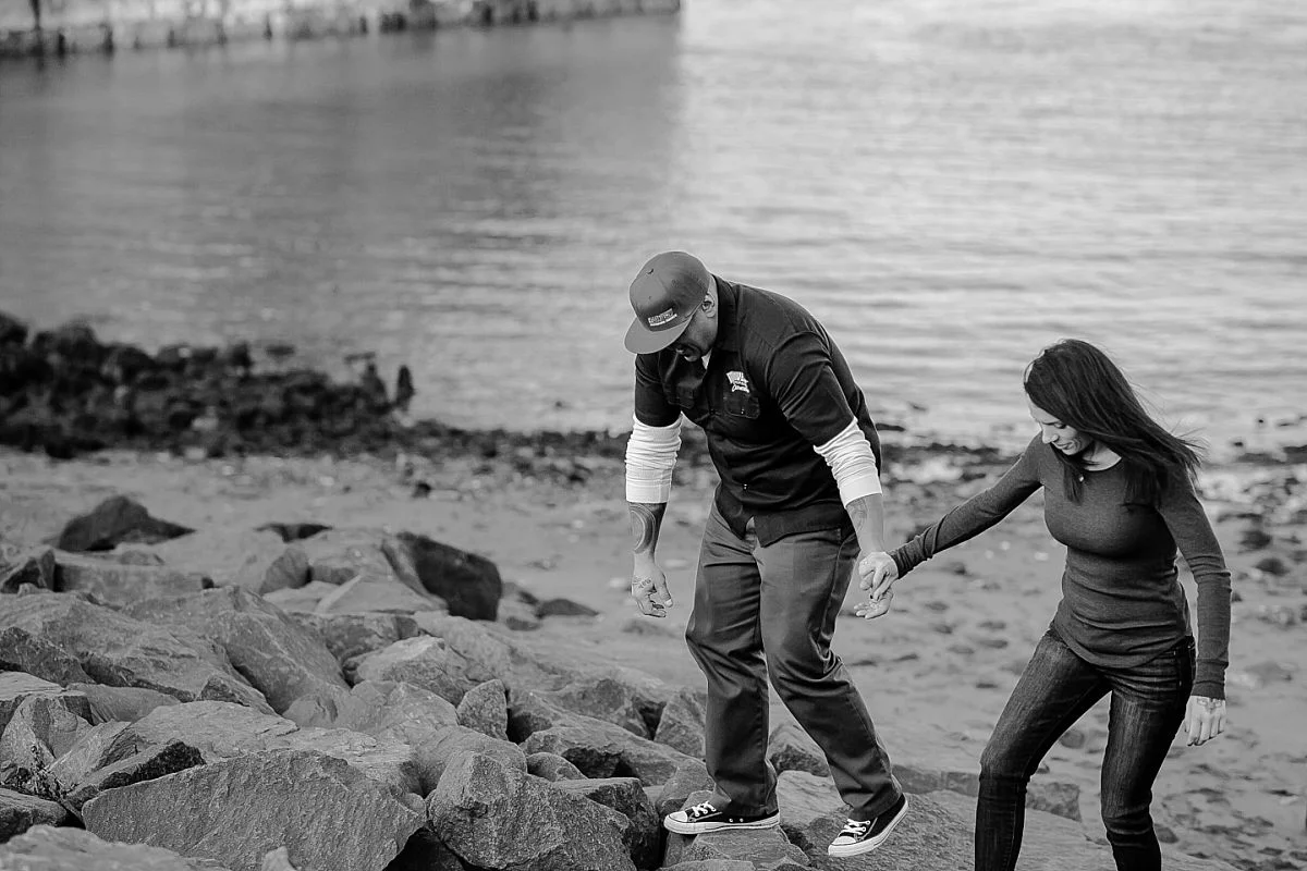 candid black and white photo of couple walking on rocks underneath the manhattan bridge in dumbo brooklyn. image by nyc family photographer krystil mcdowall