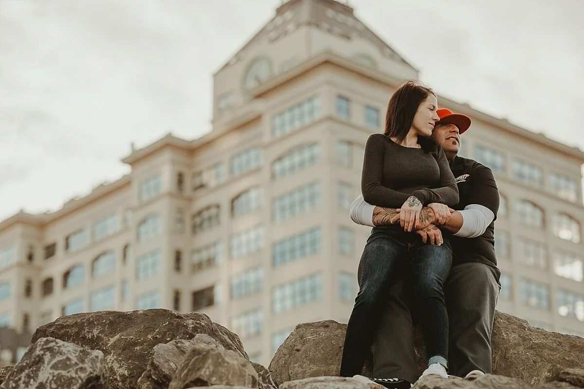couple sits on rocks in front of manhattan bridge in dumbo brooklyn while brooklyn buildings sit in the background. image by nyc family photographer krystil mcdowall