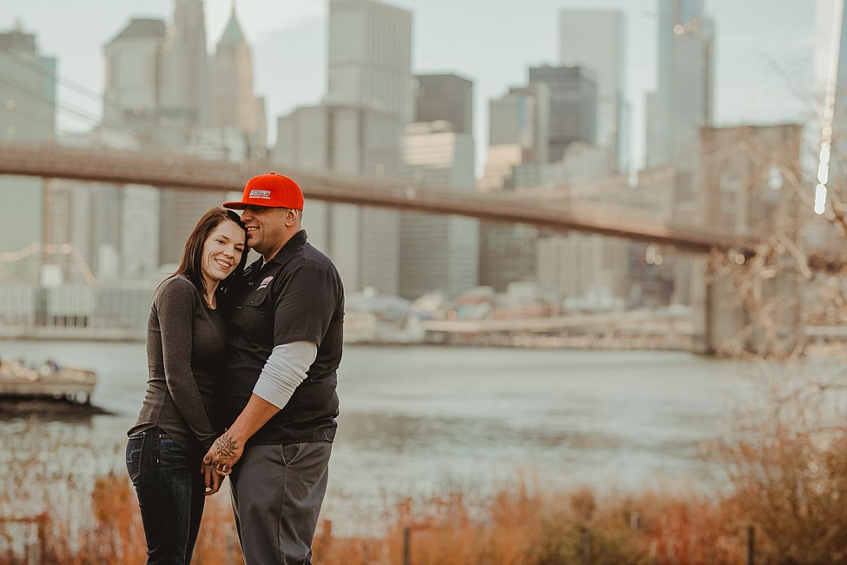 couple posed on green grass in dumbo brooklyn while beautiful manhattan skyline and brooklyn bridge appear in the background. image by nyc family photographer krystil mcdowall