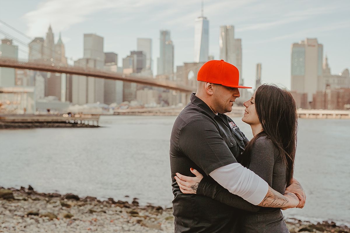 couple embraces on beach front in dumbo brooklyn looking out at beautiful manhattan skyline and brooklyn bridge. image by nyc family photographer krystil mcdowall