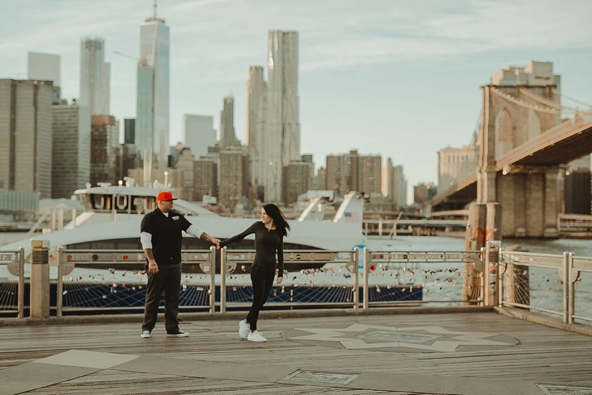 couple walks along pier in brooklyn bridge park with manhattan skyline and brooklyn bridge in the background. image by nyc family photographer krystil mcdowall