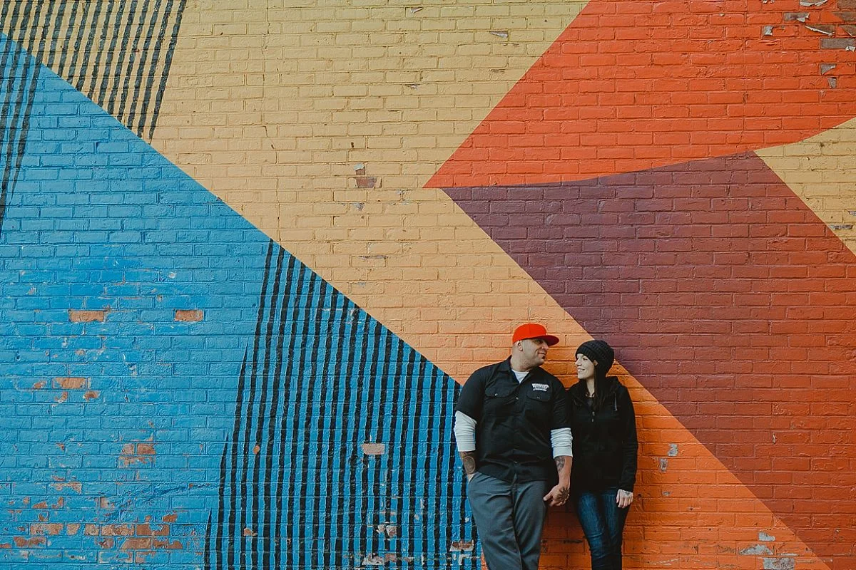couple holds hands and stands in front of colorful graffiti mural in brooklyn. photo by nyc family photographer krystil mcdowall