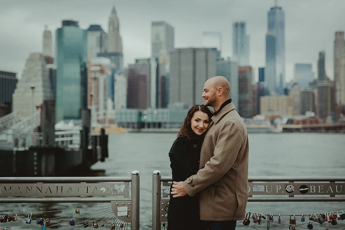 couples photo of wife looking at the camera and embracing her husband at dumbo pier with nyc skyline in the background. image by nyc family photographer krystil mcdowall