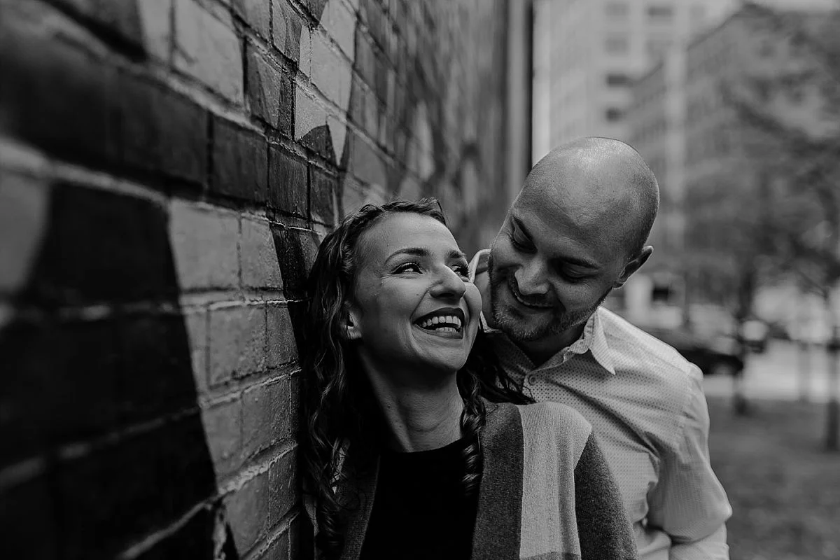 black and white photo of husband and wife giggling in front of graffiti mural in dumbo brooklyn. photo by nyc family photographer krystil mcdowall