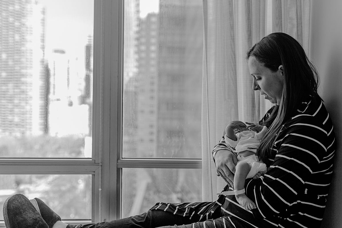 fresh 48 photo of mom holding newborn son while sitting in hospital window and looking at son lovingly. photo by nyc family and newborn photographer krystil mcdowall