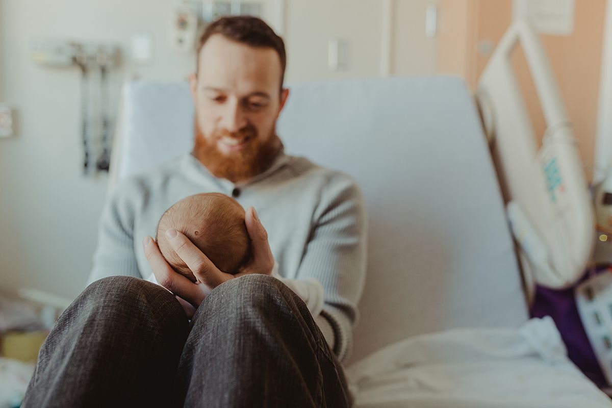 fresh 48 photo of dad sitting on hospital bed and holding newborn son in his arms with a big and loving smile. photo by nyc family and newborn photographer krystil mcdowall