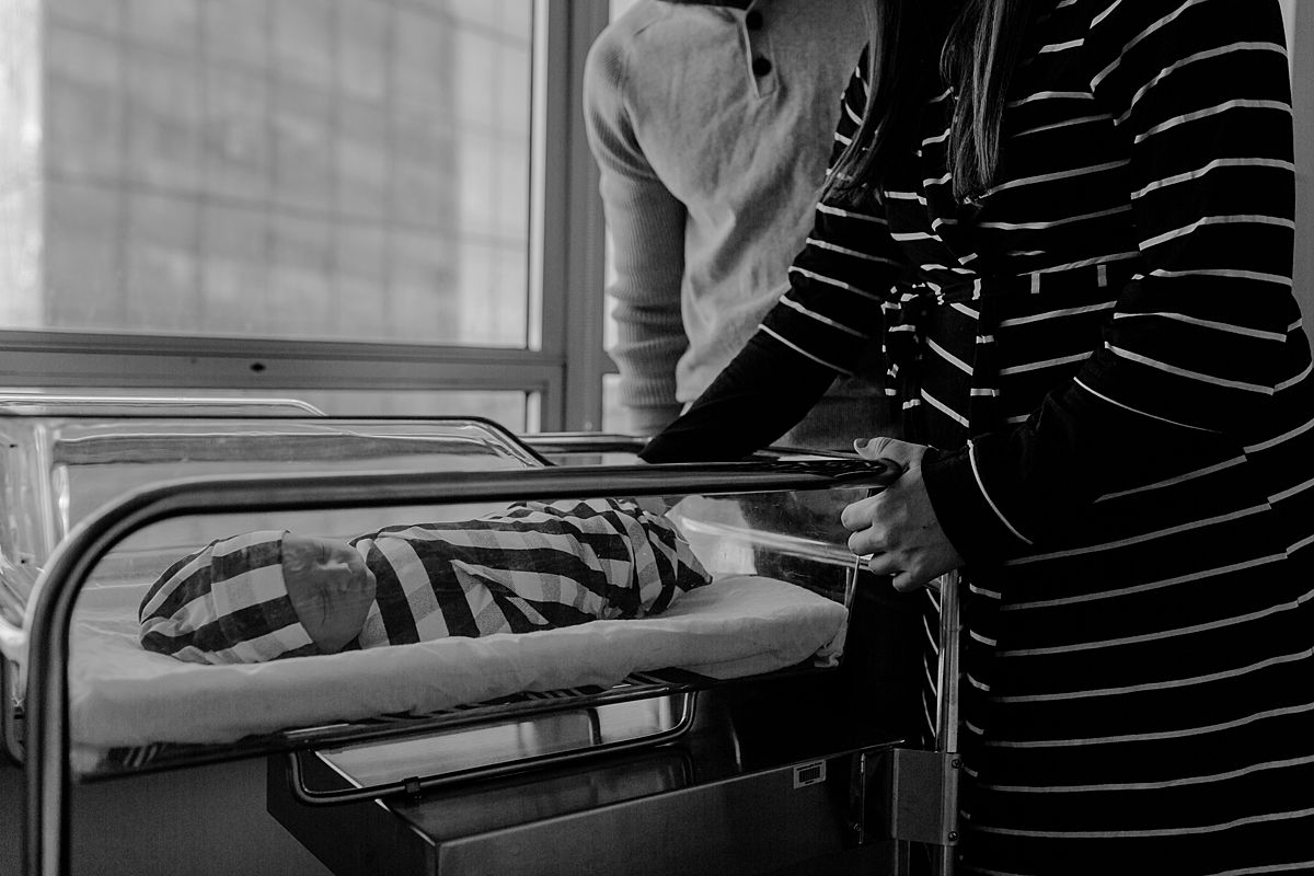 fresh 48 photo of mom and dad looking on newborn son as he sleeps in hospital bassinet. photo by nyc family and newborn photographer krystil mcdowall