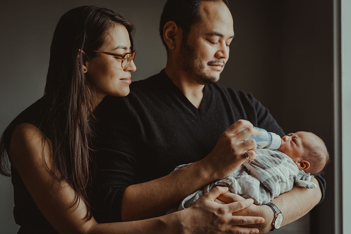 portrait of mom and dad feeding newborn baby boy near light filled window of nyc apartment. photo by nyc family and newborn photographer krystil mcdowall