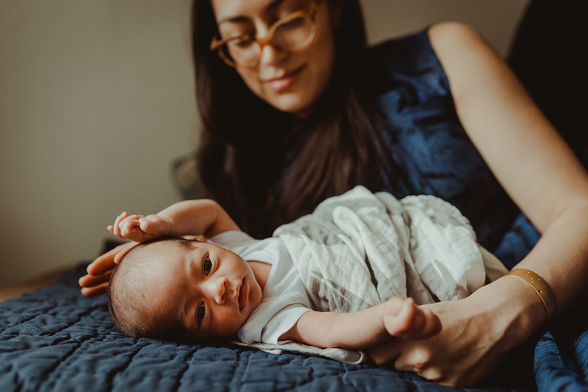 newborn son laying wrapped in whale swaddle on couch in nyc lounge room while mom lays peacefully behind him. photo by nyc family and newborn photographer krystil mcdowall