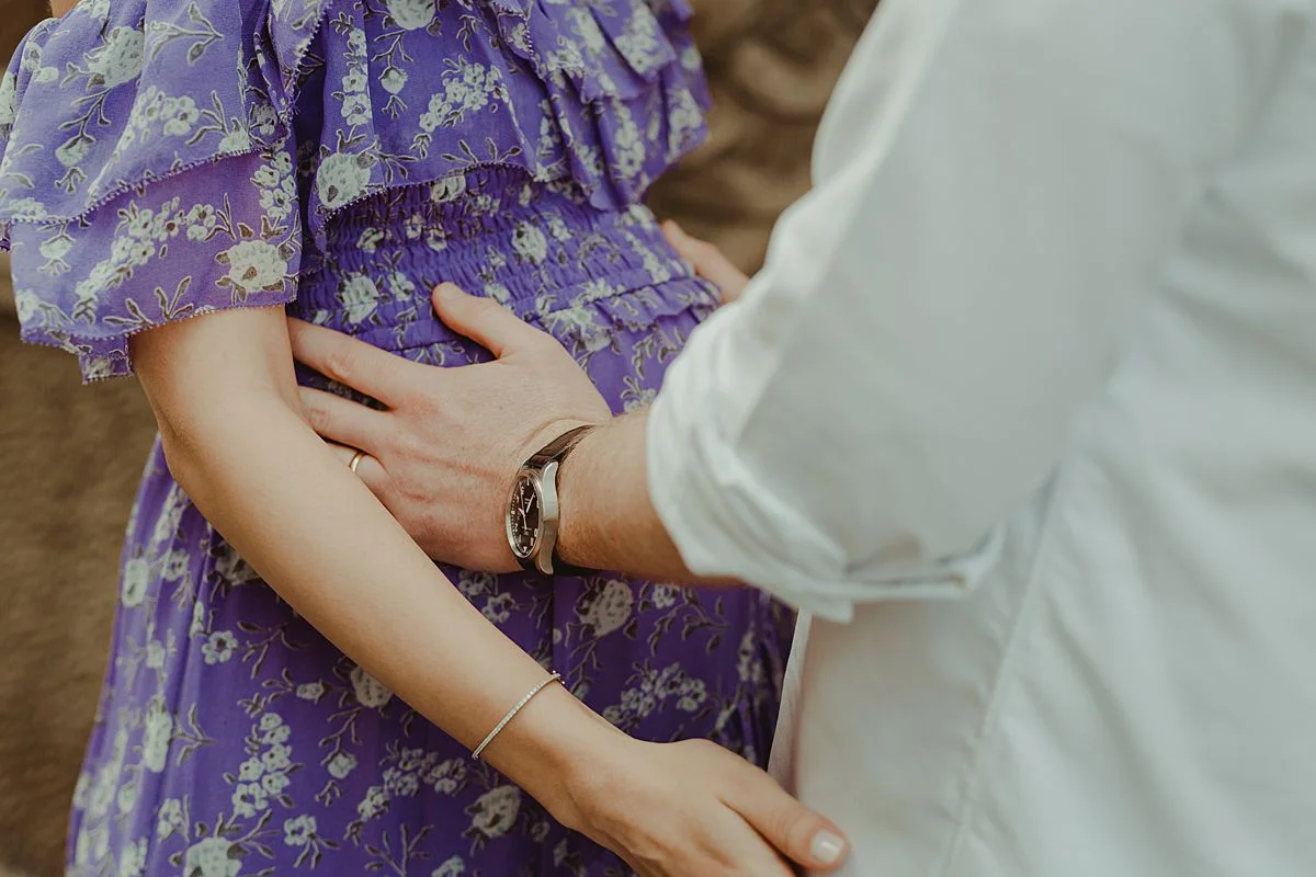 close up shot of expecting dad holding expecting mom's pregnant belly. mom dressed in feminine purple and white flower dress. maternity photo by nyc family and newborn photographer krystil mcdowall
