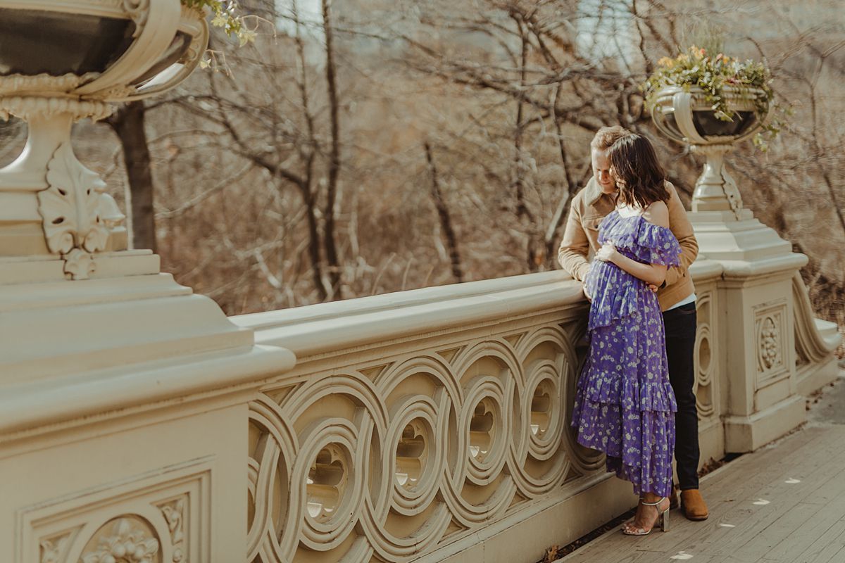 expecting couple stands on bow bridge in central park looking out at nyc skyline during maternity photo session. image by nyc family and newborn photographer krystil mcdowall