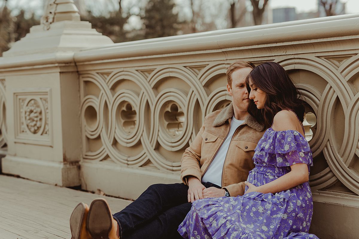 expecting couple sits on bow bridge in central park holding mama's expecting belly during maternity photo session. image by nyc family and newborn photographer krystil mcdowall