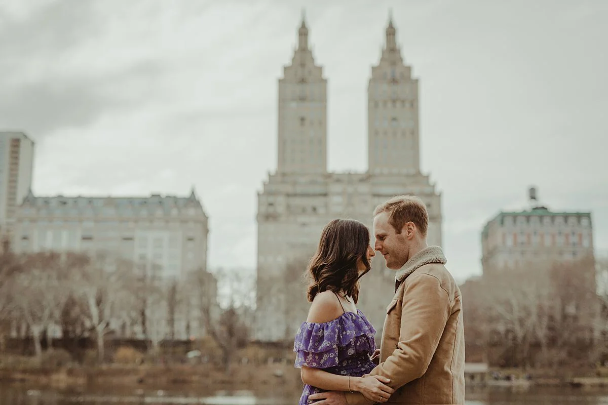 expecting couple stands in front of lake in central park with the the san remo building in the background. image by nyc family and newborn photographer krystil mcdowall