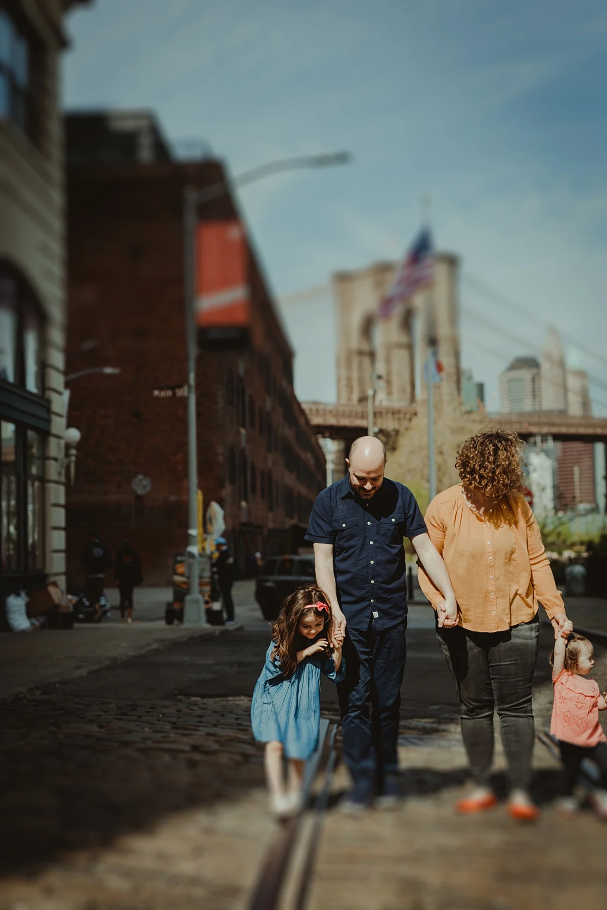 tilt shift candid family photo of family walking along cobble stoned street in dumbo brooklyn. photo by nyc family and newborn photographer krystil mcdowall
