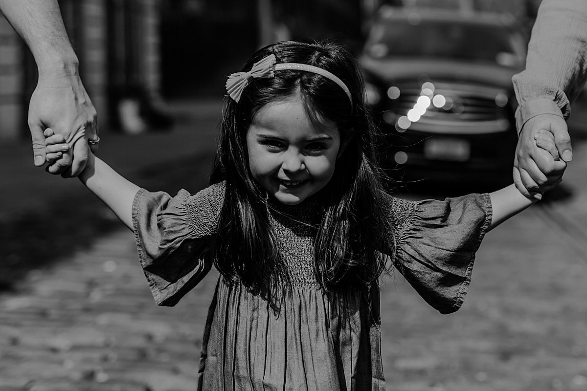 photo of young girl holding mom and dad's hands as she walks along cobble stoned street in dumbo brooklyn. photo by krystil mcdowall photography