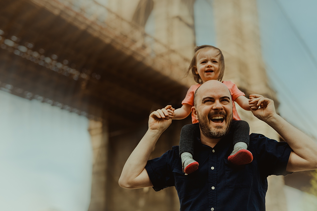 photo of dad giving young daughter a shoulder ride with blurred brooklyn bridge in the background. photo by nyc family and newborn photographer krystil mcdowall