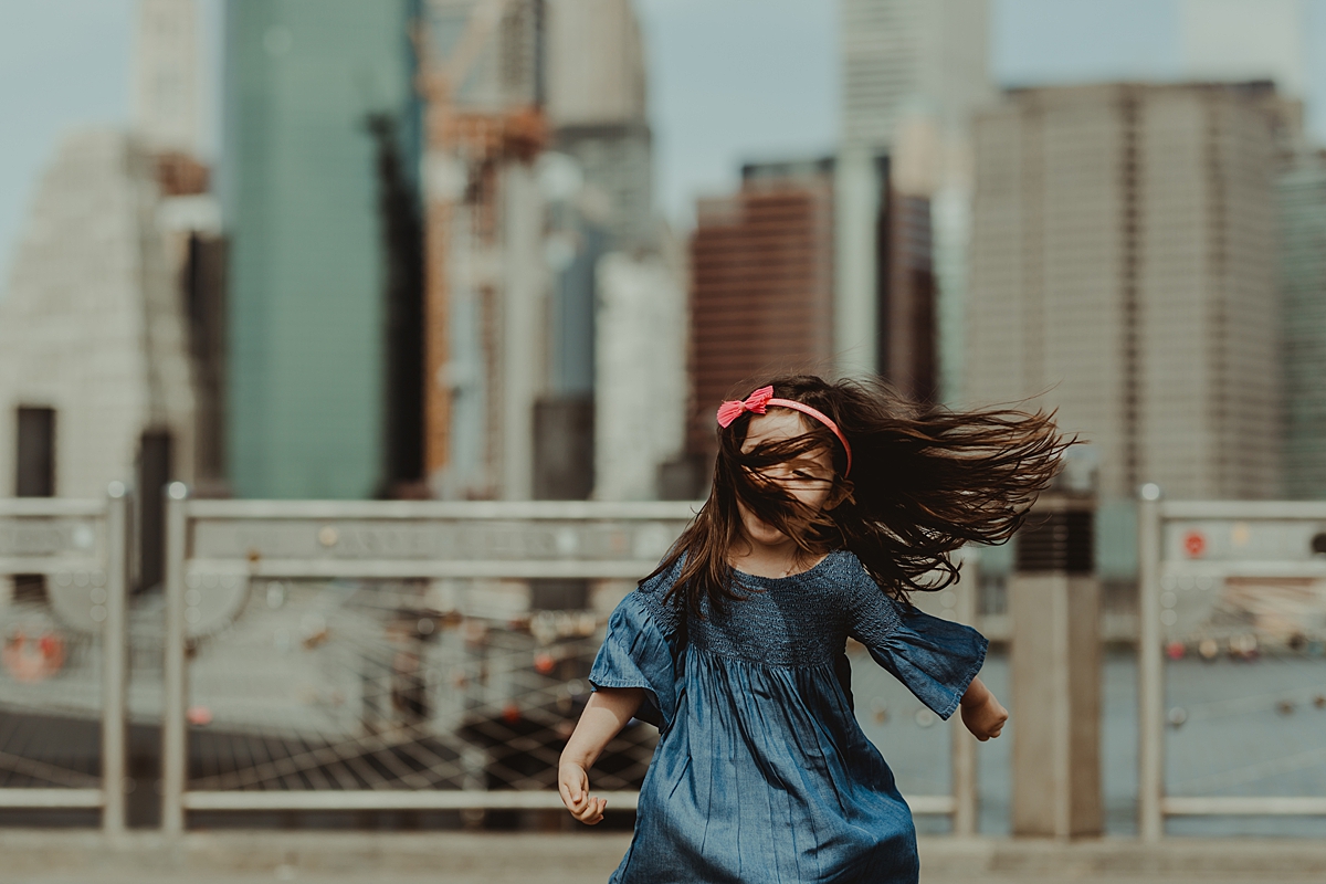 candid portrait of young daughter in blue dress playing at brooklyn bridge park in front of love locks with manhattan skyline in the background. photo by nyc family and newborn photographer krystil mcdowall