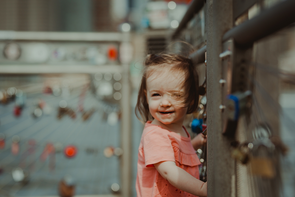 candid portrait of daughter in pink shirt standing in front of love locks with manhattan in the background. photo by nyc family and newborn photographer krystil mcdowall