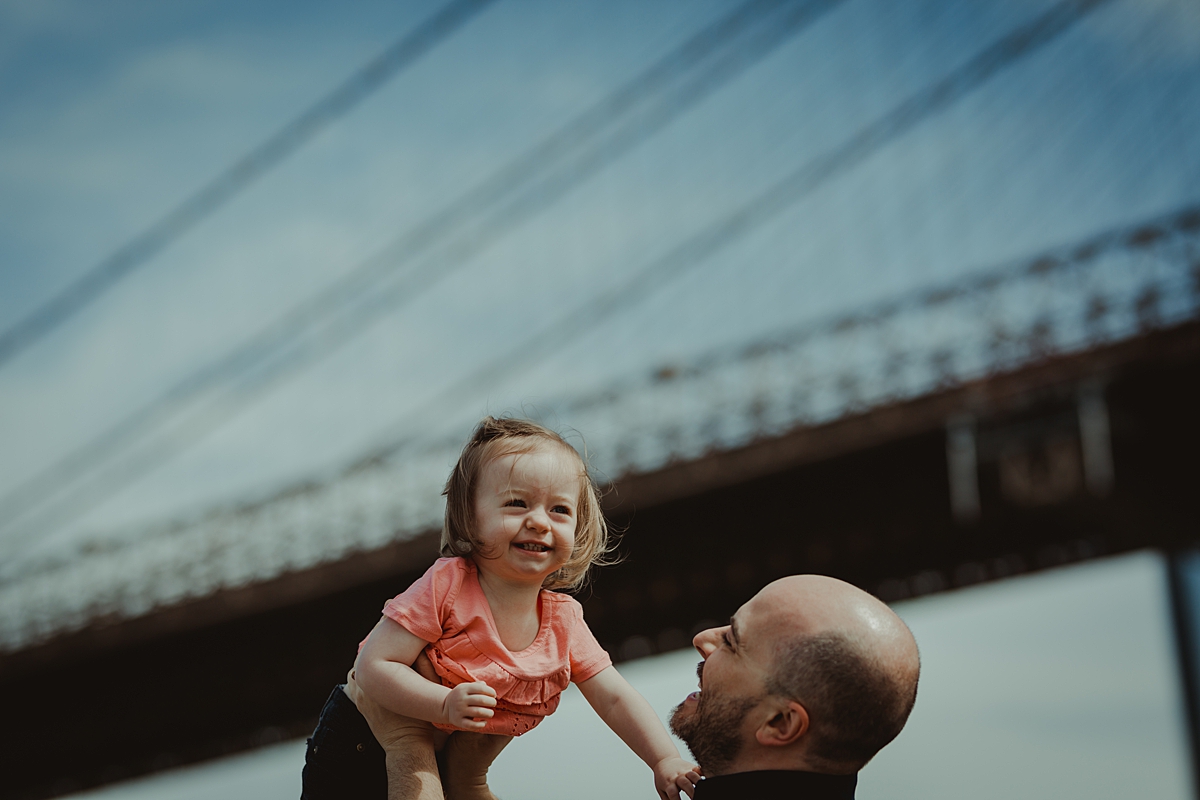 dad holding laughing daughter in the air in front of the brooklyn bridge in dumbo. photo by nyc family and newborn photographer krystil mcdowall