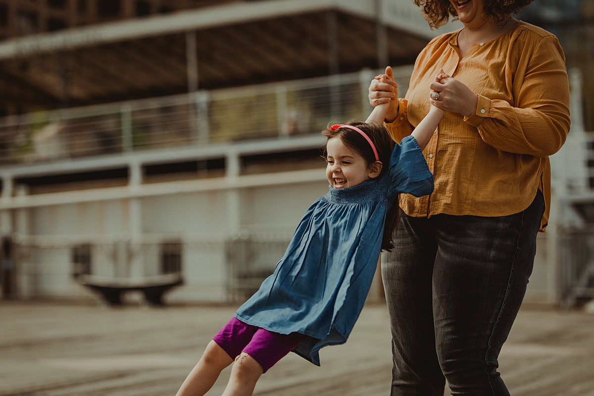 candid photo of mom spinning daughter around while she's laughing at the pier at brooklyn bridge park. photo by nyc family and newborn photographer krystil mcdowall