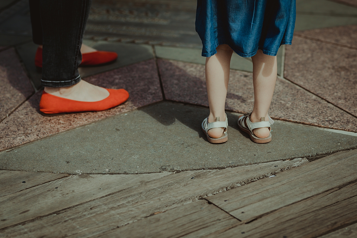 detail photo of mom's bright red flats and daughter's white buckle up shoes taken while standing at the pier at brooklyn bridge park. photo by nyc family and newborn photographer krystil mcdowall