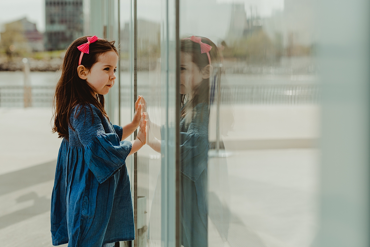 photo of young girl in blue dress with pink headband standing in front of jane's carousel in dumbo brooklyn and looking in at carousel with reflection in the glass. photo by nyc family and newborn photographer krystil mcdowall
