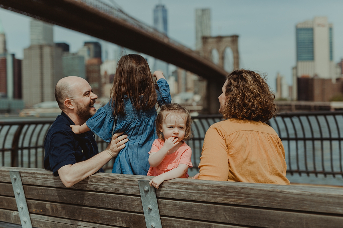 photo of family sitting on park bench with brooklyn bridge in the background. photo by nyc family and newborn photographer krystil mcdowall
