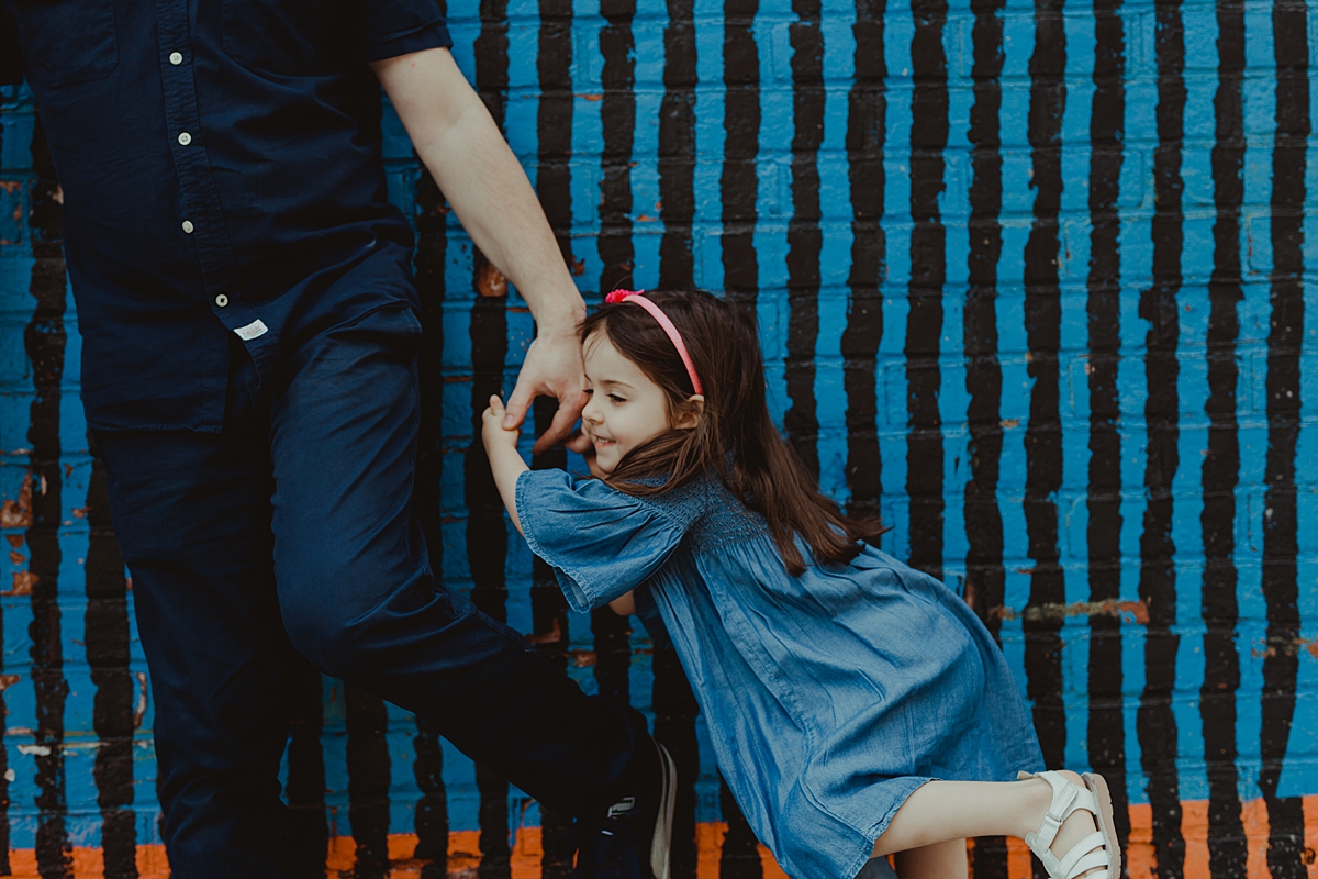 photo of daughter holding dad's hand and standing in front of bright blue and black graffiti wall in dumbo brooklyn. photo by nyc family and newborn photographer krystil mcdowall